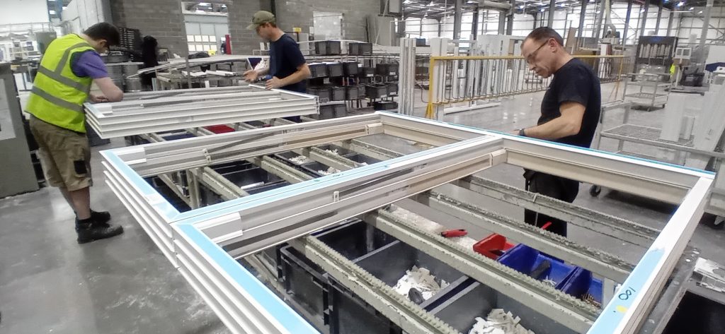 Two men assembling a large metal frame in the Victorian Sliders factory, focused on their work in a bustling environment.